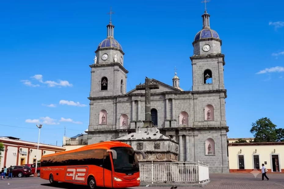 Un autobús de color naranja vibrante de la compañía Rapi2s, con su logo visible, estacionado en la plaza adoquinada frente a la histórica Catedral de Ciudad Guzmán bajo un cielo azul claro.