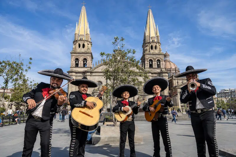 Grupo de cinco músicos de mariachi con traje tradicional negro y sombrero de charro tocando instrumentos frente a la Catedral de Guadalajara.