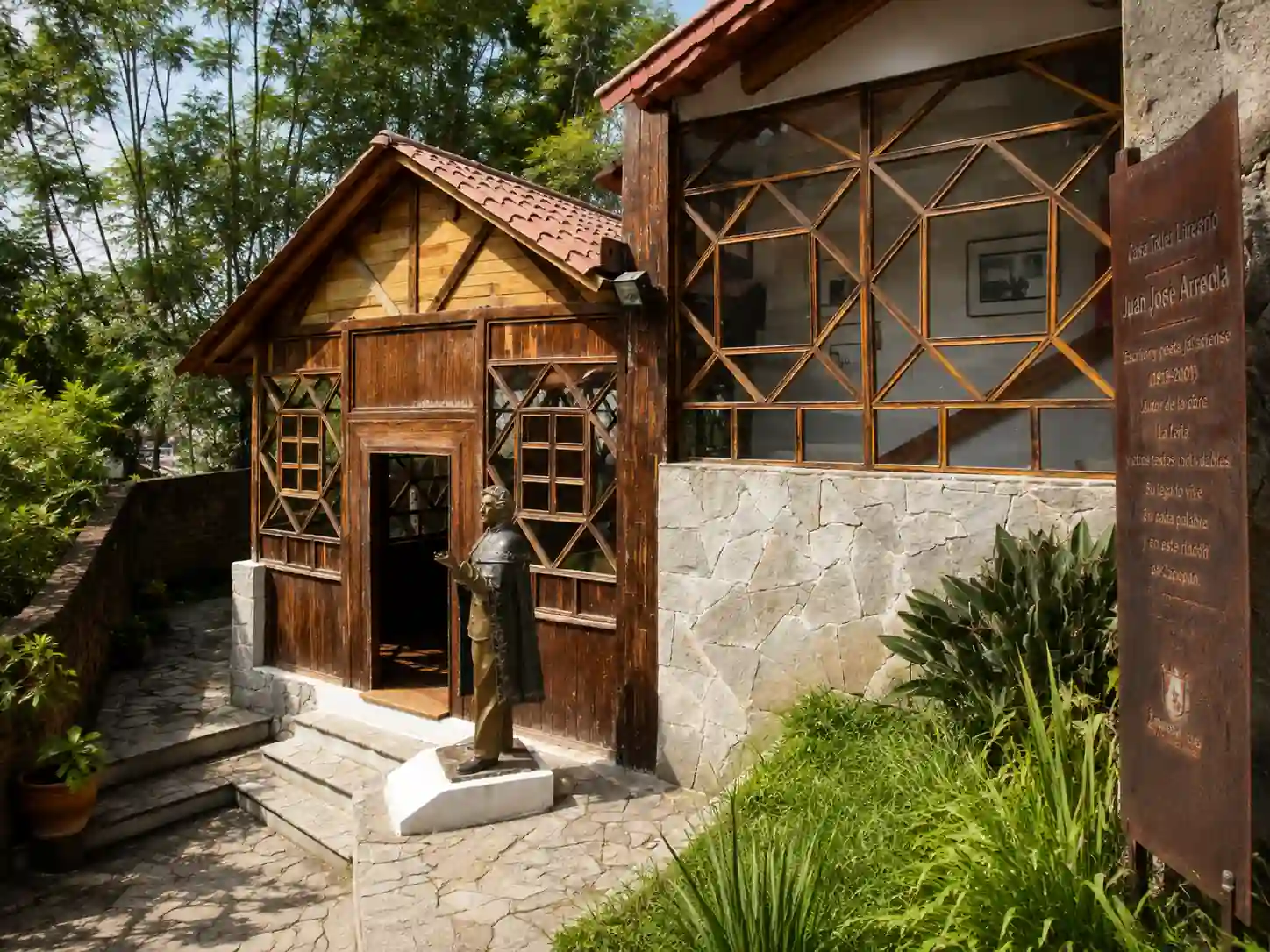 Fachada de la Casa Taller Literario Juan José Arreola con estructura de madera, una estatua de bronce al frente y un letrero informativo lateral.