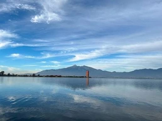 Vista panorámica de la Laguna de Zapotlán con el volcán al fondo y su reflejo cristalino bajo un cielo con nubes dispersas.