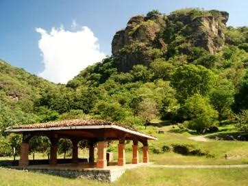 Kiosco de madera con techo de teja en un parque ecológico rodeado de áreas verdes, árboles frondosos y una imponente formación rocosa.