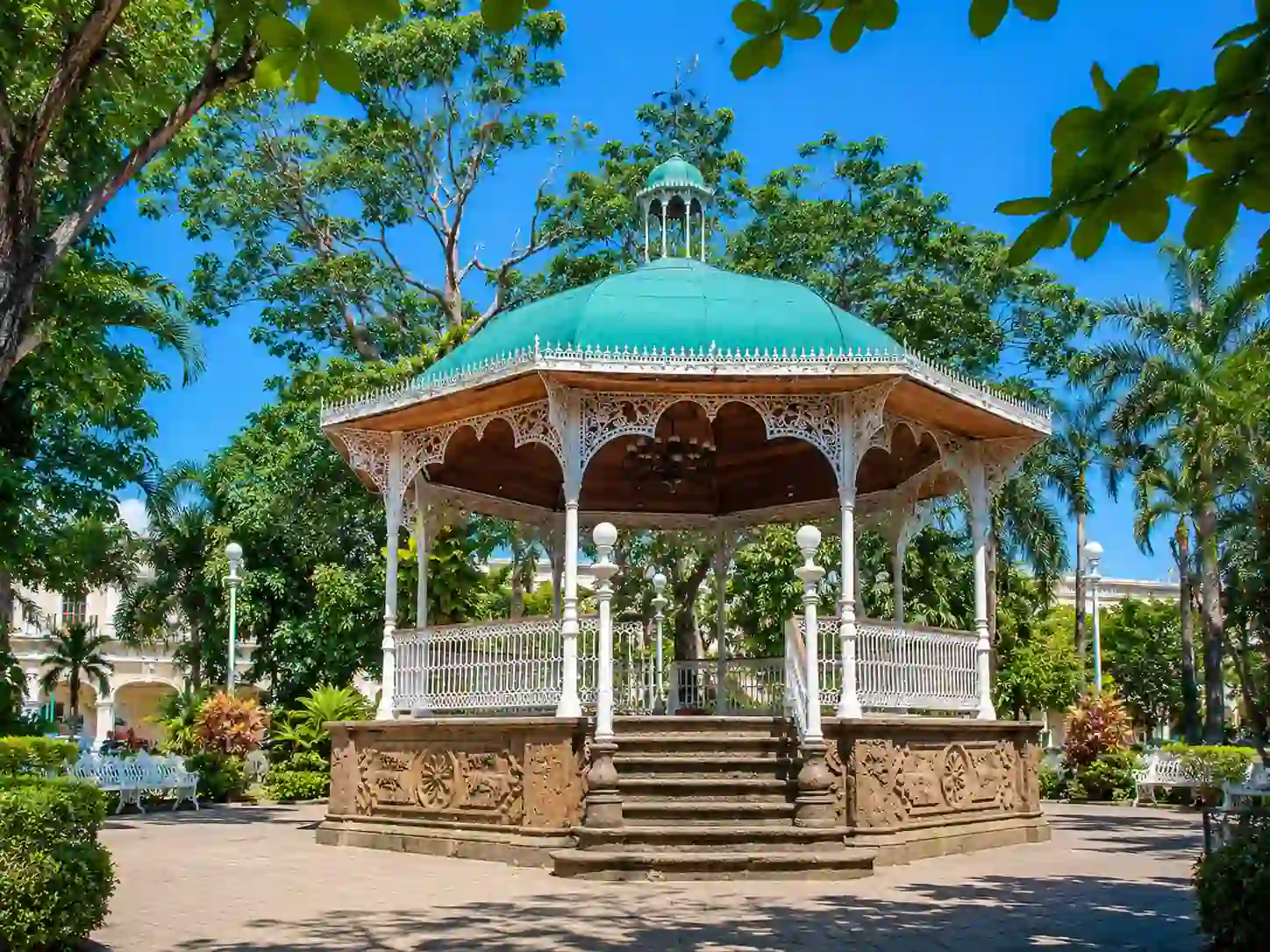 Kiosco de hierro forjado con cúpula verde y base de cantera ubicado en el Jardín Libertad de Colima, rodeado de árboles y vegetación.