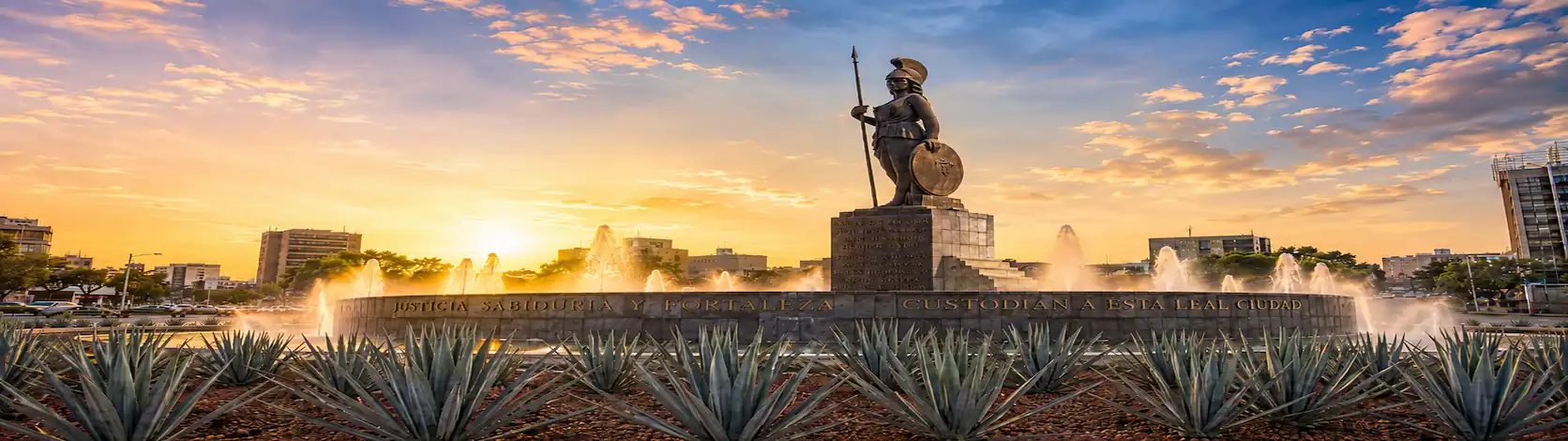 Monumento de la Minerva con fuente y esculturas rodeado de agaves bajo un cielo atardecer en Guadalajara.
