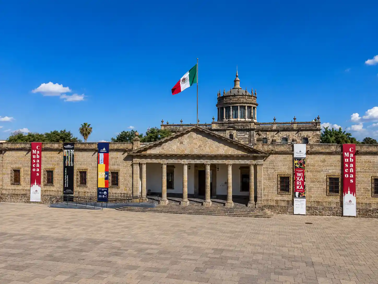 Fachada de piedra del Museo Cabañas en Guadalajara con columnas neoclásicas, la bandera de México y estandartes informativos.
