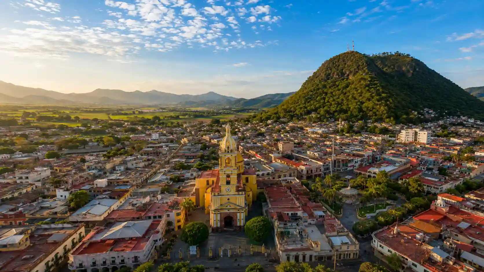 Vista aérea panorámica al atardecer de una localidad con una iglesia de cúpula amarilla, rodeada de valles y una gran montaña verde al fondo.