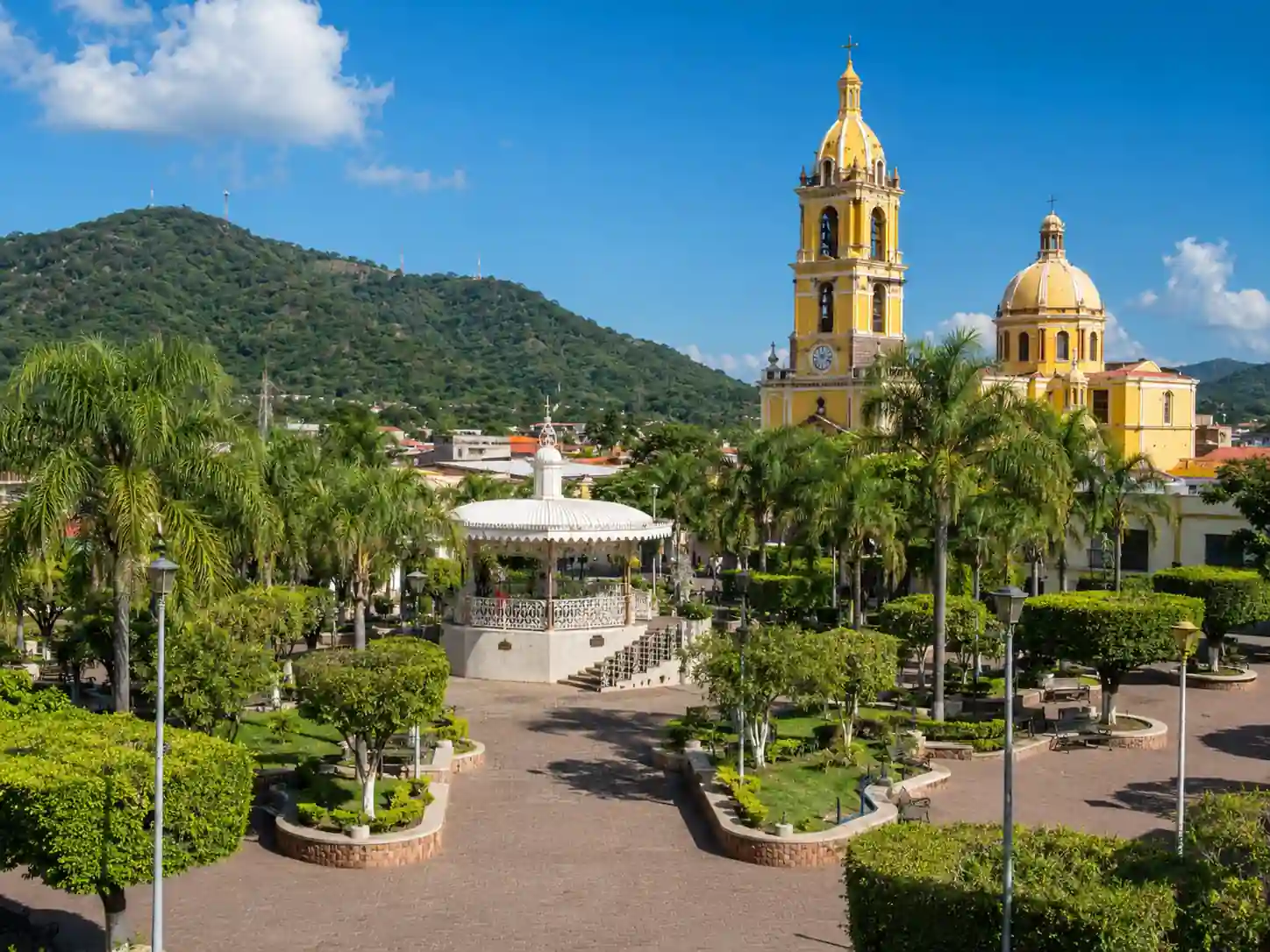 Plaza principal con un kiosco blanco de estilo tradicional, rodeada de jardines bien cuidados, palmeras y la iglesia principal con cúpula amarilla al fondo.