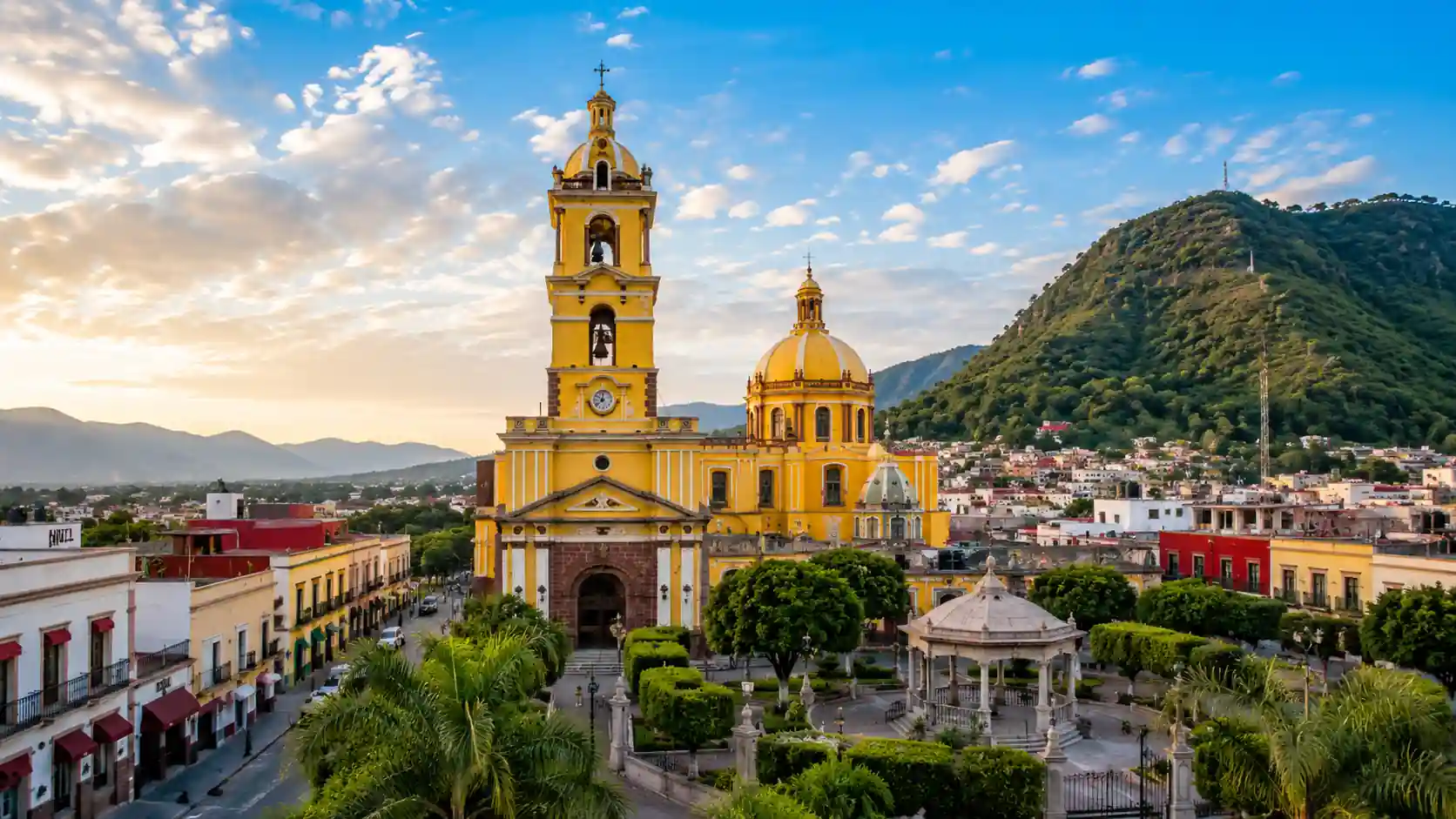 Fachada y torre campanario de un templo de color amarillo con cúpula, situado frente a una plaza con un kiosco y vegetación bajo un cielo nublado.