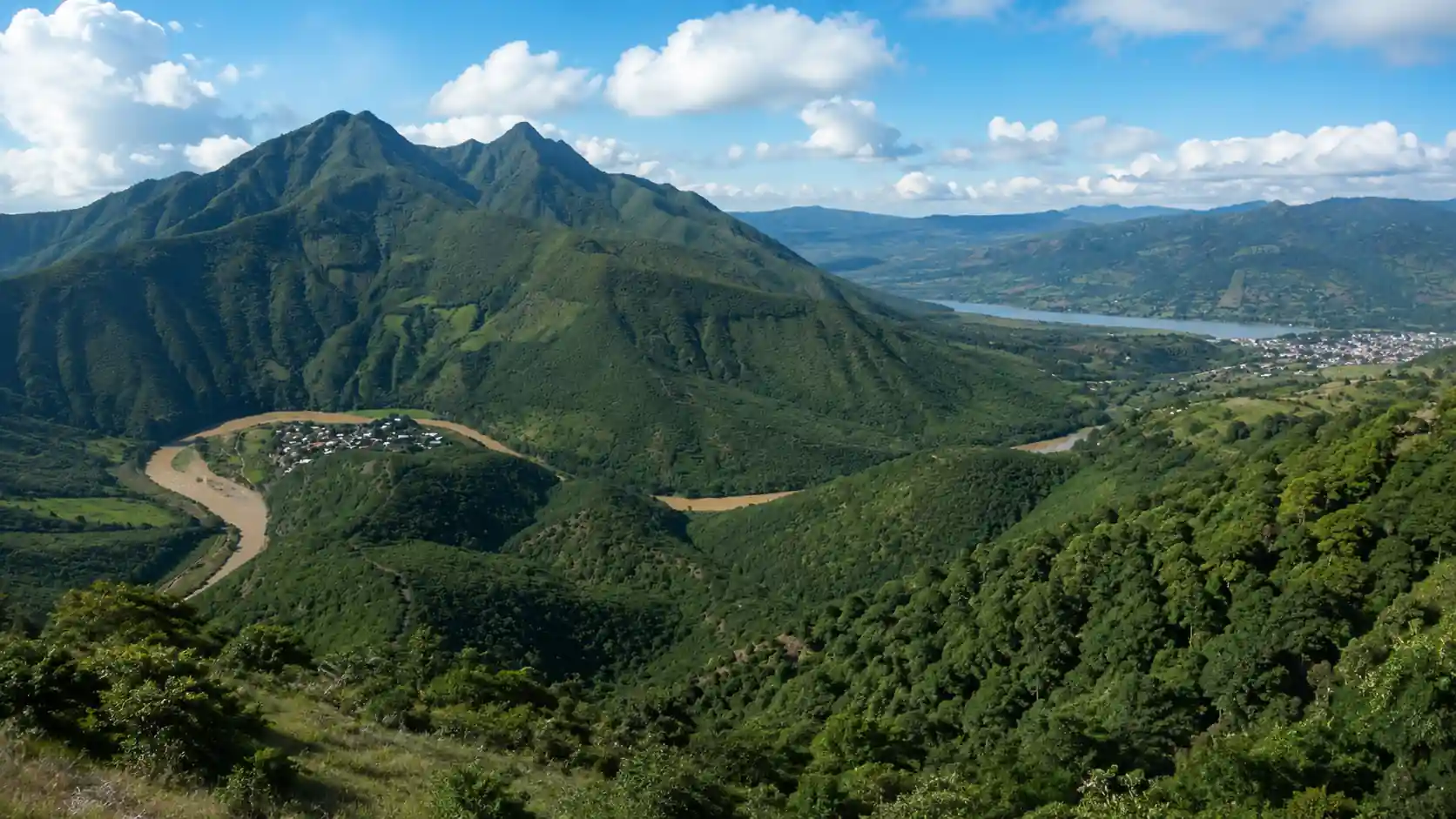 Paisaje montañoso de la Sierra del Tigre con frondosos bosques verdes, un río serpenteante y un cuerpo de agua a lo lejos bajo un cielo con nubes.