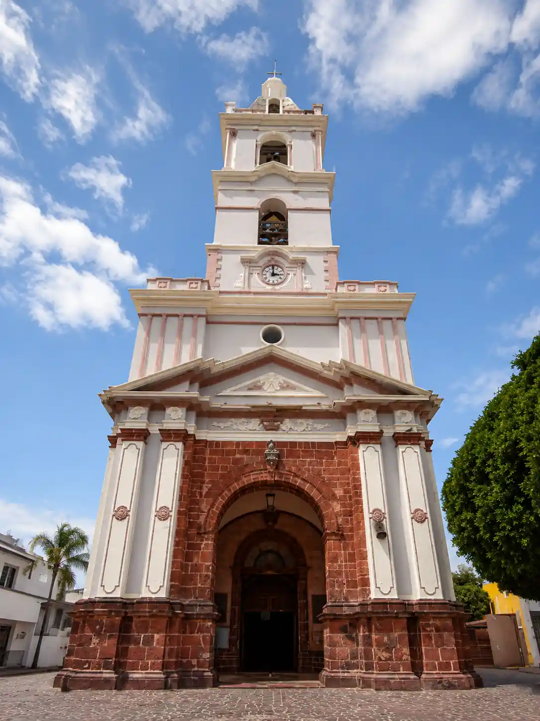 Fachada frontal de un templo religioso con base de cantera roja, columnas blancas, un arco de entrada y una torre campanario con reloj.