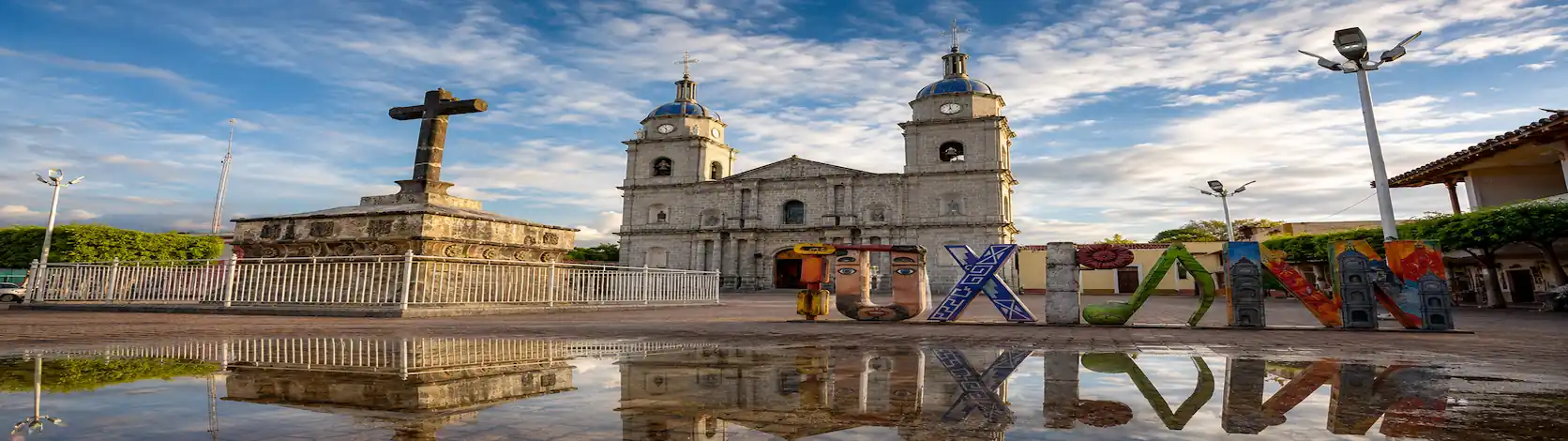 Fachada de la parroquia de Tuxpan frente a una gran cruz atrial de piedra y letras monumentales coloridas que reflejan en el suelo mojado.