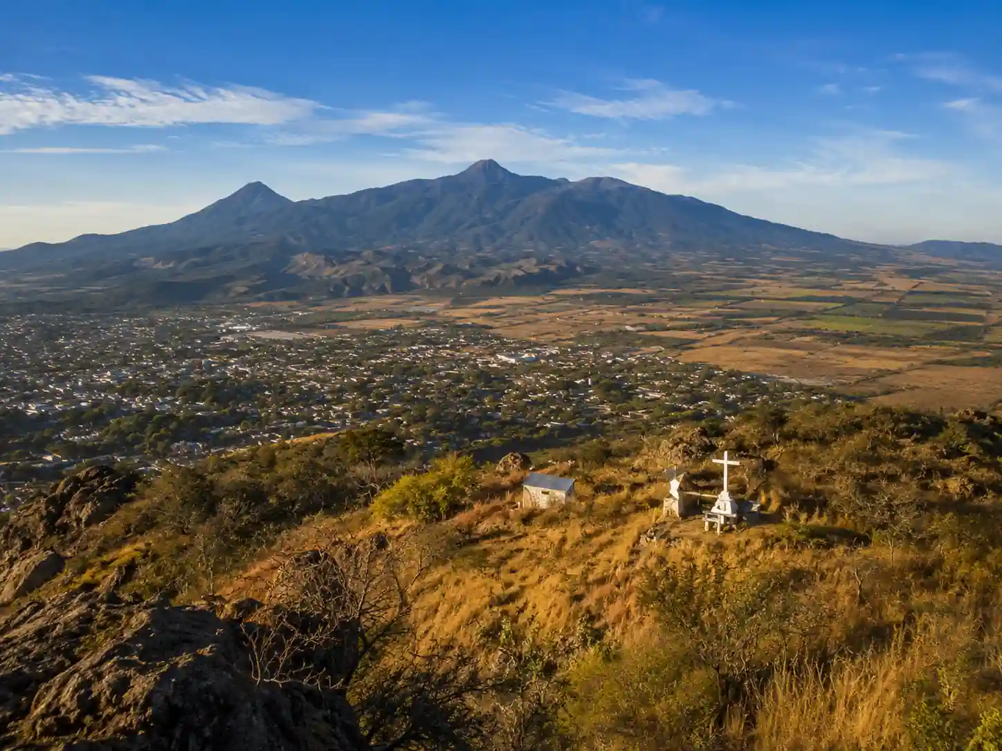 Vista panorámica desde la cima del cerro Cihuapilli con una cruz blanca, mostrando un extenso valle sembrado y montañas al fondo.