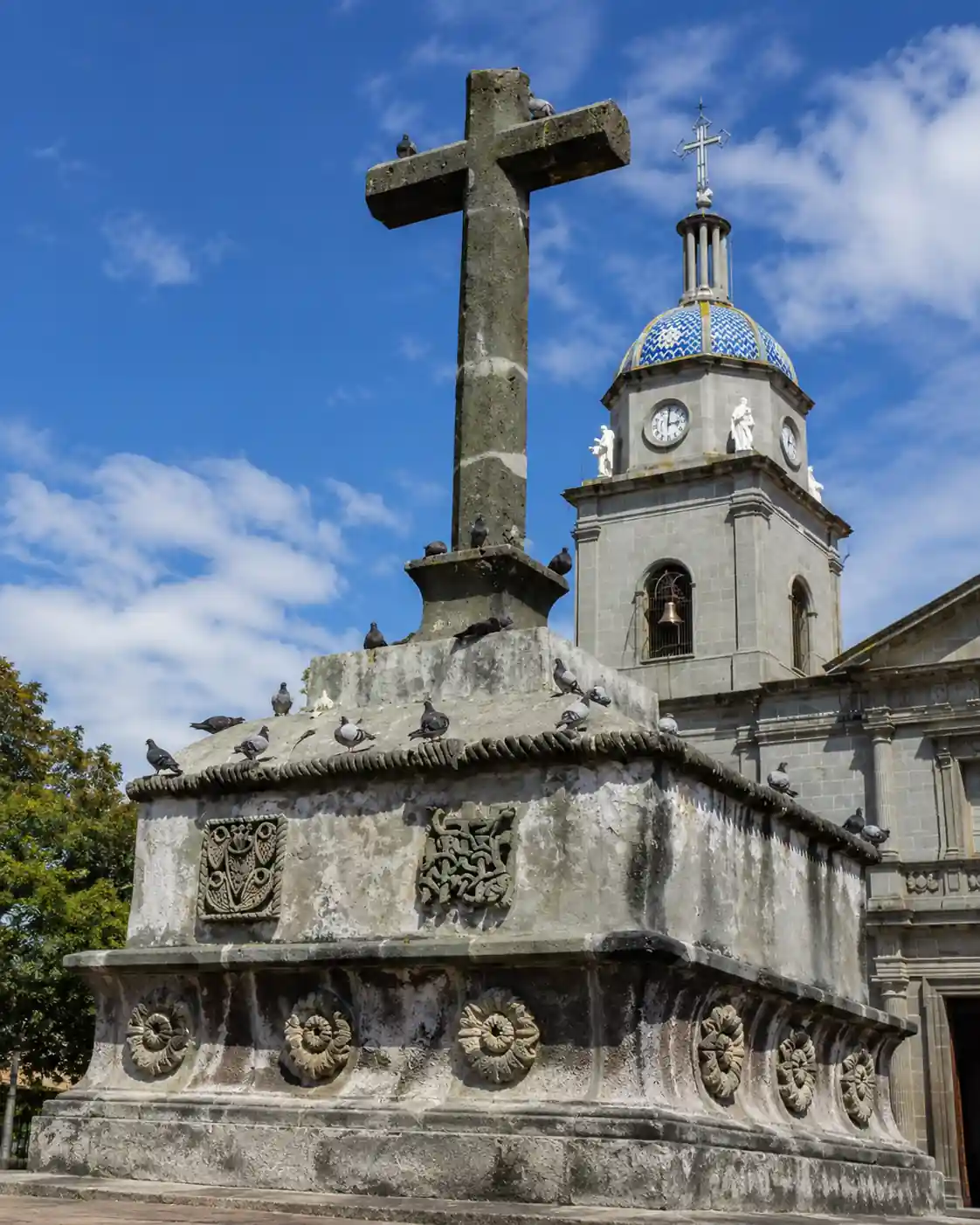 Antigua cruz atrial de piedra tallada sobre un basamento decorado con relieves y palomas posadas, con la torre de una iglesia de cúpula azul al fondo.