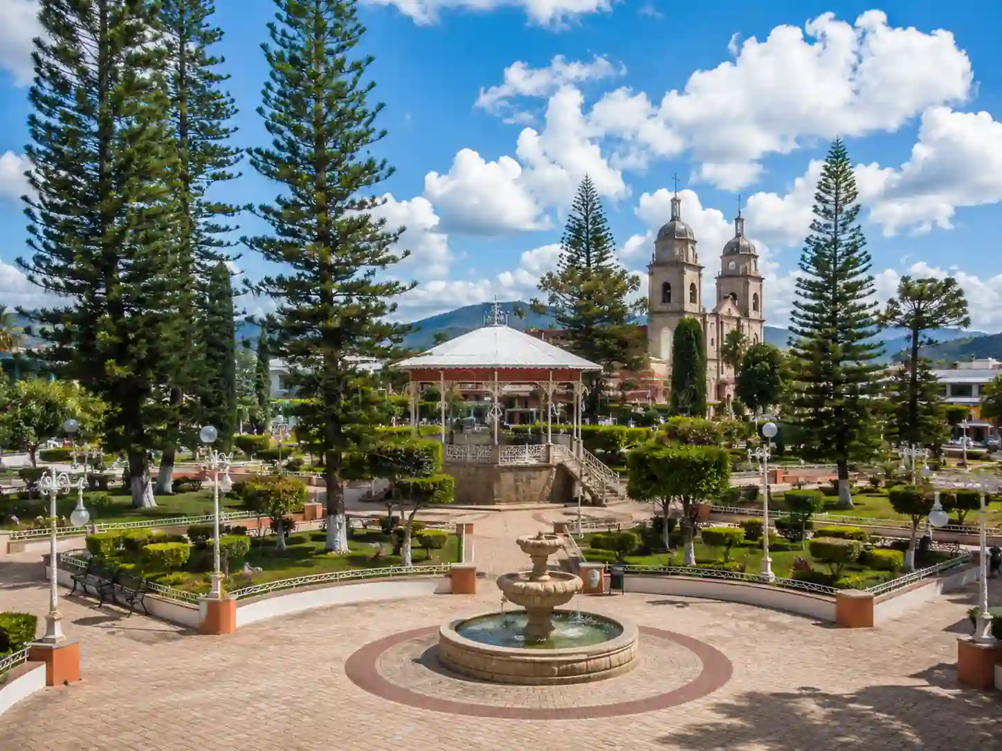Jardín municipal con árboles altos, un kiosco blanco de estilo tradicional y una fuente de piedra en primer plano bajo un cielo nublado.