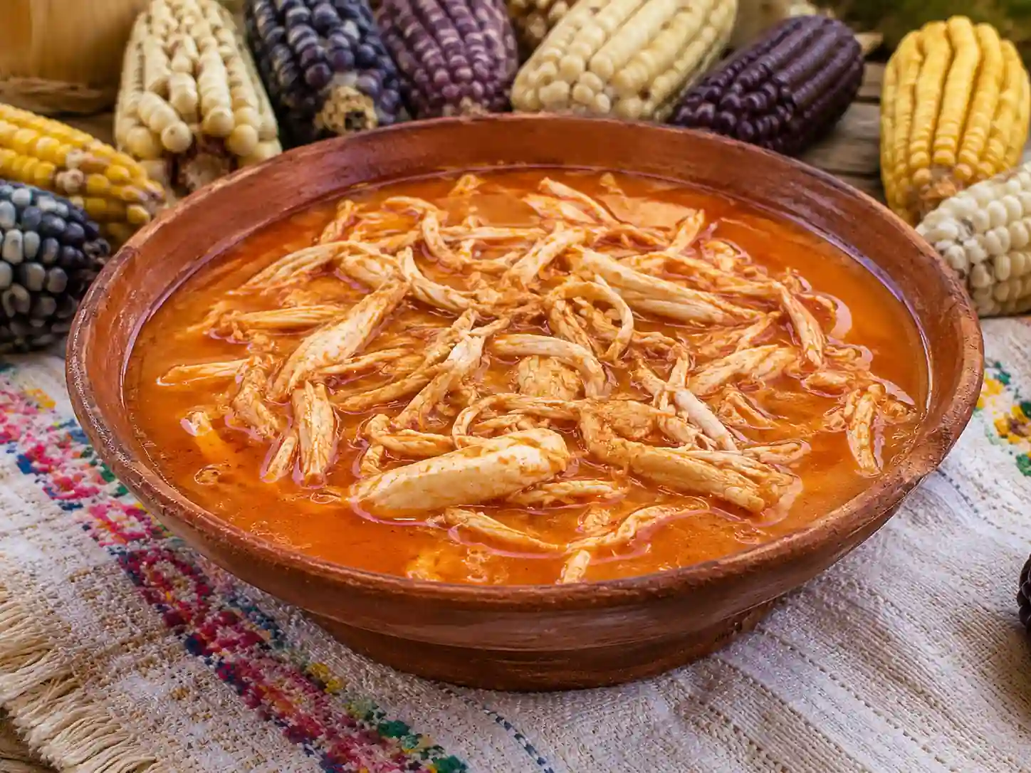 Plato de barro con caldo de coaxala rojo con pollo deshebrado, rodeado de coloridas mazorcas de maíz nativo sobre un mantel tradicional.