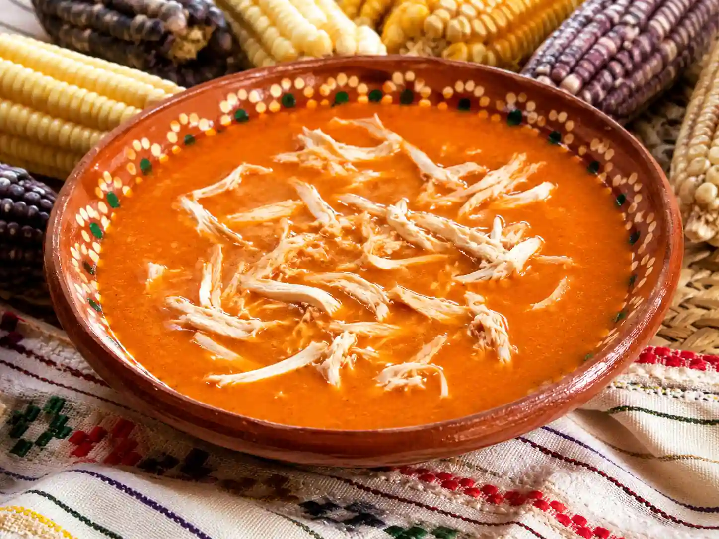Plato de barro tradicional con cuachala de pollo en salsa espesa roja, rodeado de mazorcas de maíz de diferentes colores.