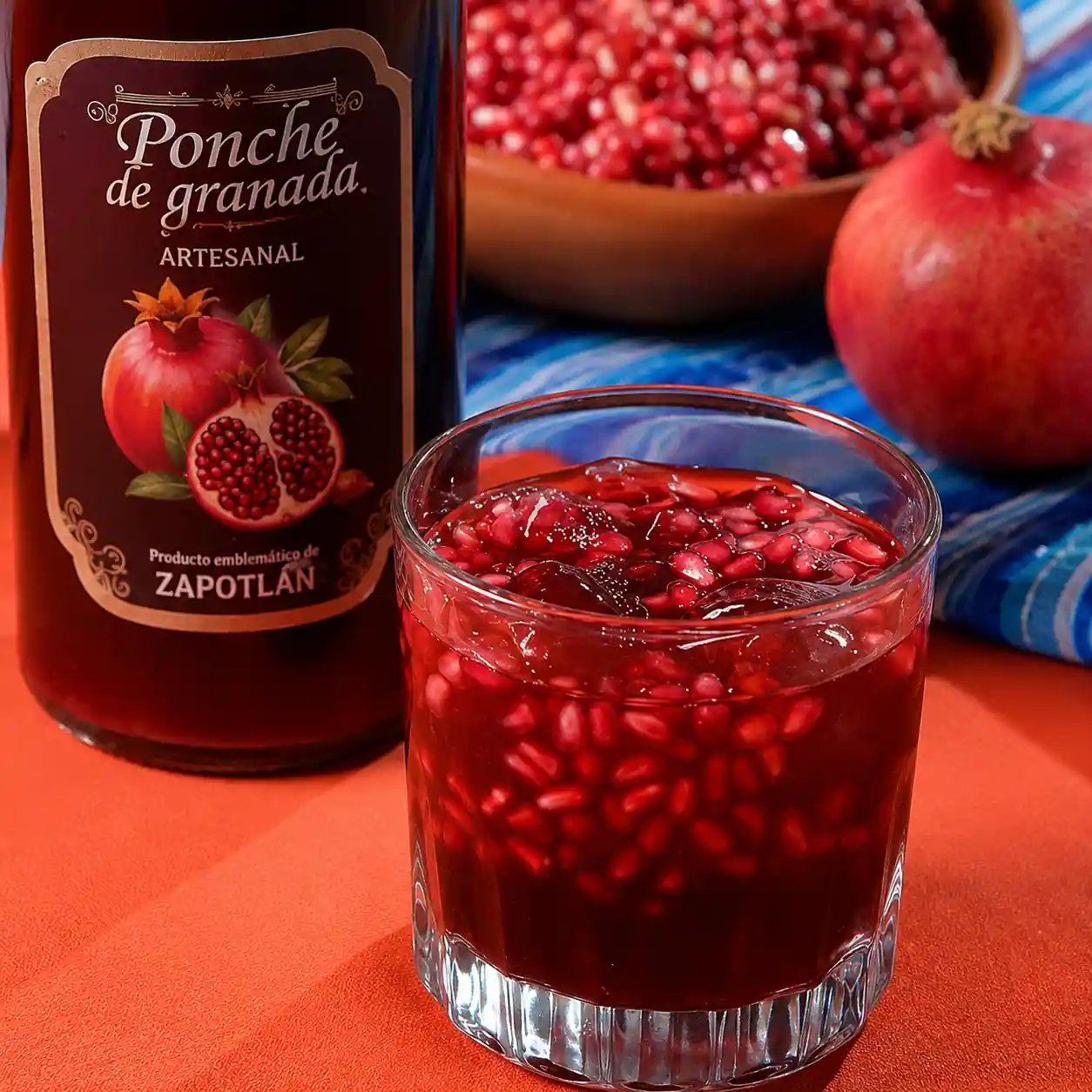 Vaso de ponche artesanal de granada con granos frescos y hielos, junto a una botella rotulada y granadas enteras sobre la mesa.